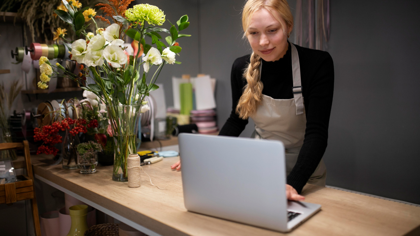 A young female stands at a table next to some flowers, looking at her laptop and discovering the new TransferGo Business multi-currency account.