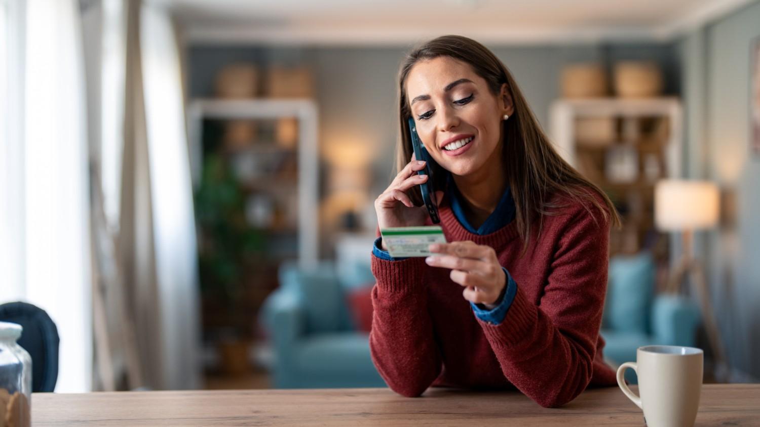 A young female sits at a desk whilst holding her bank card and speaking on her smartphone to ask the question 'Is Revolut Business safe?'