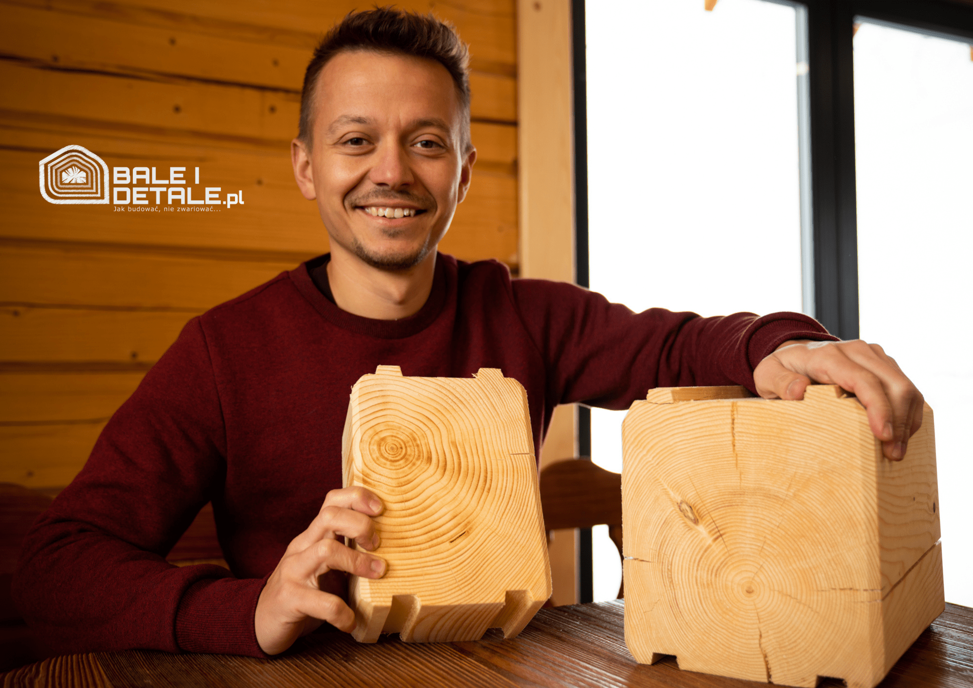 A young man, who is the founder of BaleiDetale.pl, smiles as he stands in a red jumper holding some chopped wood