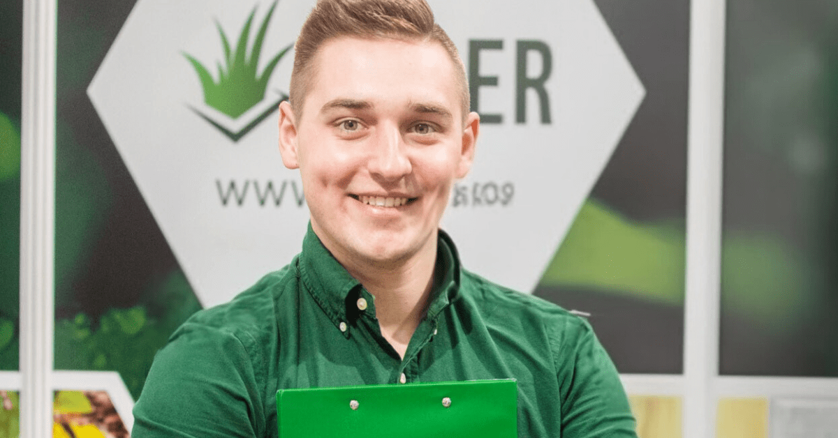 A young male in a green uniform smiles as he stands in front of an Edger business sign